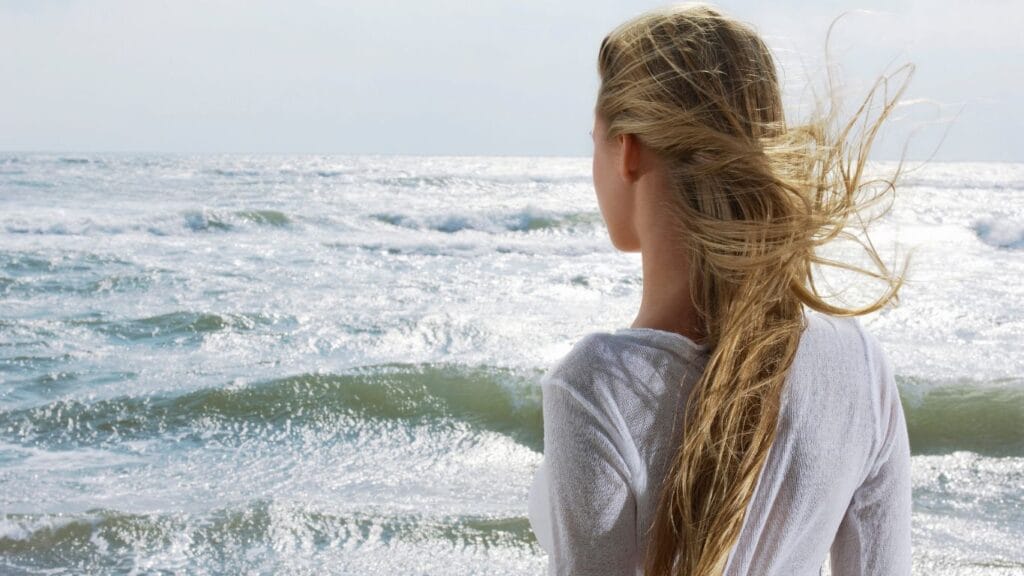 Woman with long blonde hair blowing in ocean wind standing at beach — natural hair care routine to protect hair from saltwater sun and wind damage