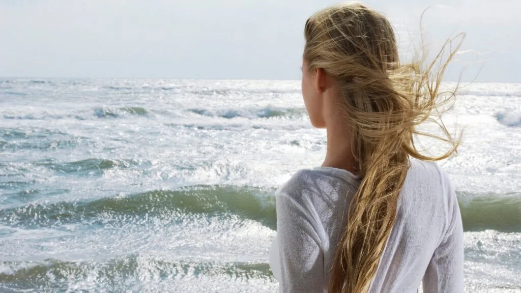 Woman with long blonde hair blowing in ocean wind standing at beach — natural hair care routine to protect hair from saltwater sun and wind damage