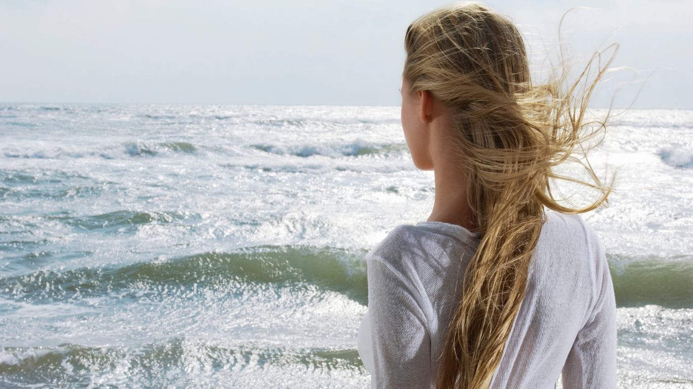 Woman with long blonde hair blowing in ocean wind standing at beach — natural hair care routine to protect hair from saltwater sun and wind damage